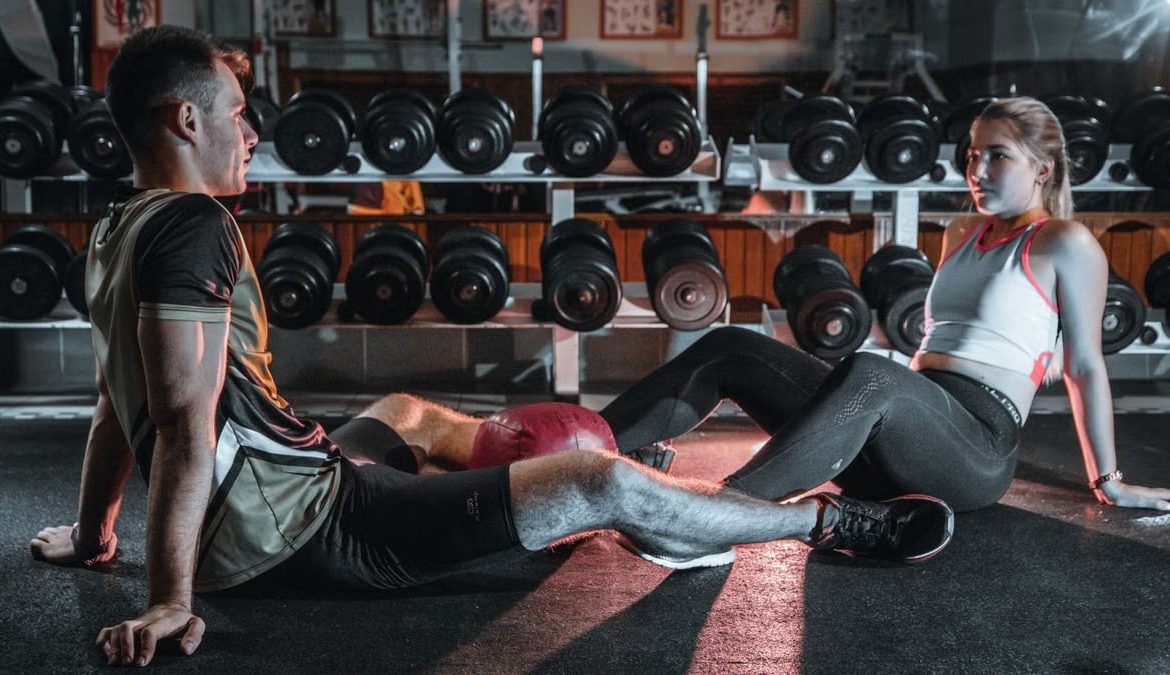 A man and woman resting together on the gym floor, surrounded by dumbbells.